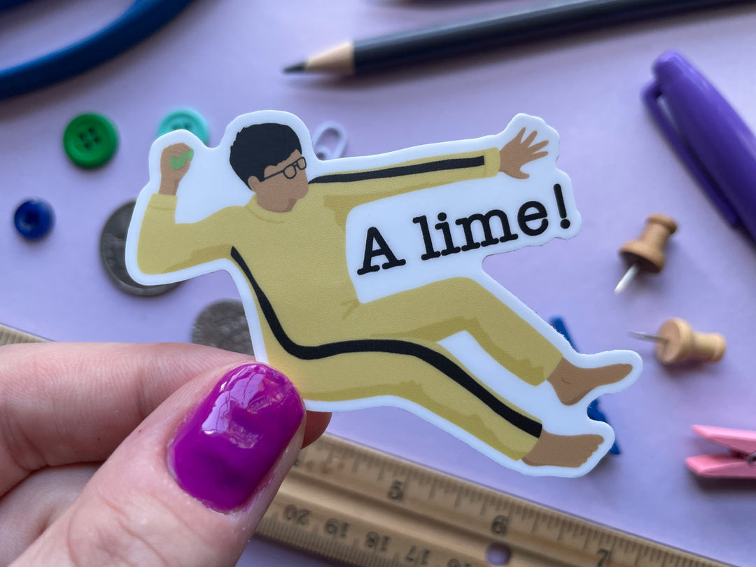 A sticker of comedian Phil Wang in a yellow jumpsuit with a black stripe sitting on the floor, barefoot, throwing a lime with the text &quot;A lime!&quot; is shown held above a purple background.