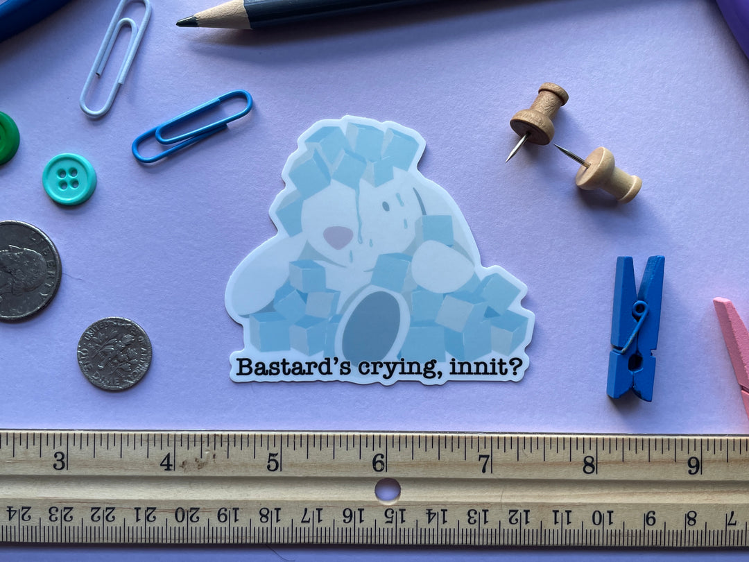 A sticker from the TV show Taskmaster of a stuffed rabbit covered in blue ice cubes and water. Under the rabbit is the text &quot;Bastard&#39;s crying, innit?&quot; The sticker is shown on a purple background surrounded by small objects.