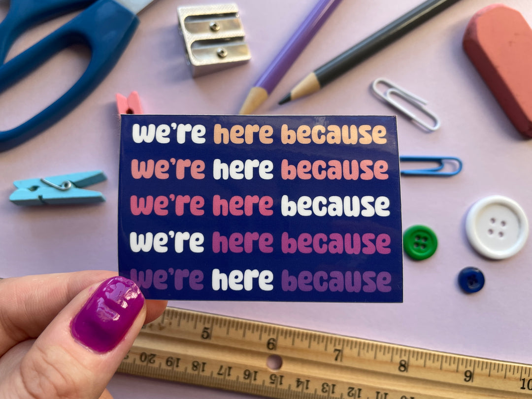 A dark blue sticker with colorful text in bubble letters reads "we're here because" repeating five times. The sticker is held above a purple background covered in objects.