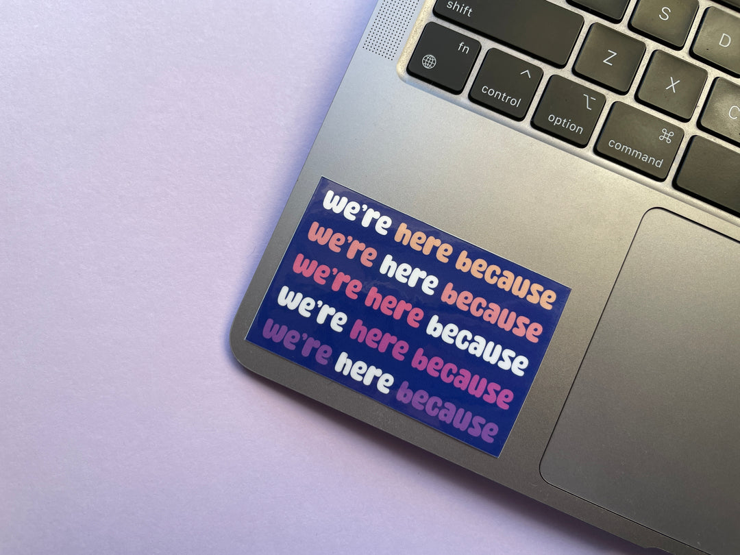 A dark blue sticker with colorful text in bubble letters reads "we're here because" repeating five times. The sticker is on silver laptop on a purple background covered in objects.