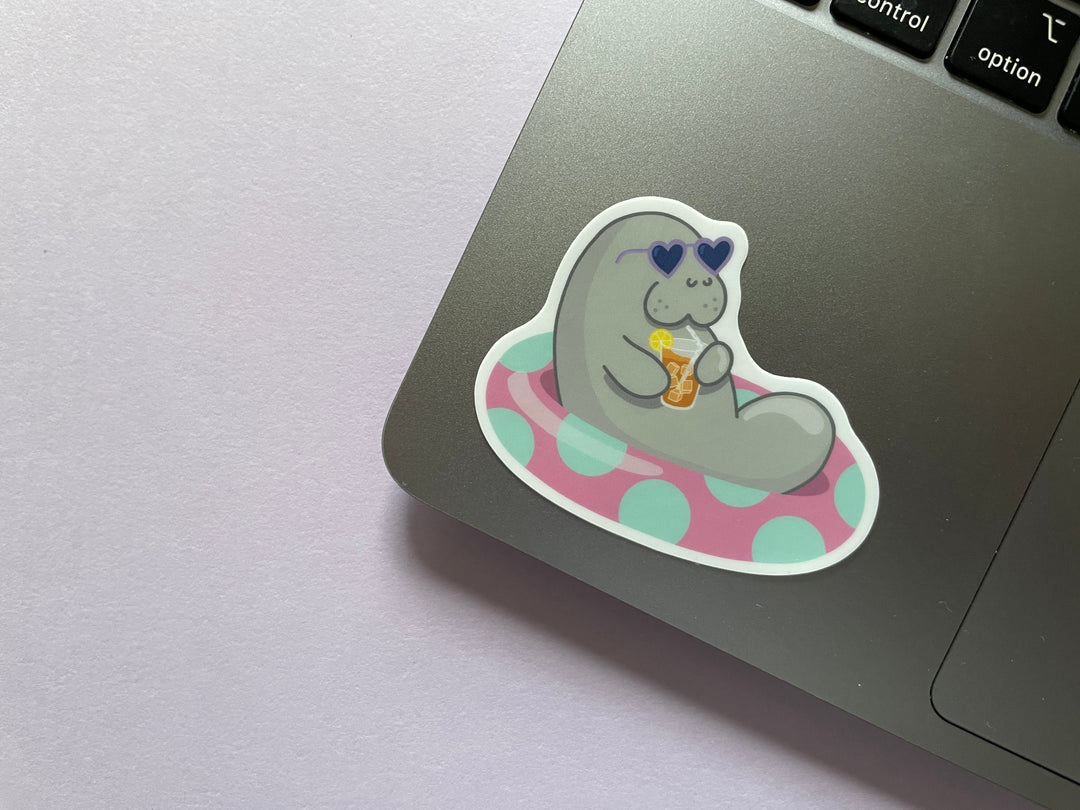 A sticker of a grey cartoon manatee with purple heart-shaped sunglasses holding a glass of iced tea lounging in a pink inner tube with green polka dots is shown on a silver laptop on a purple background.