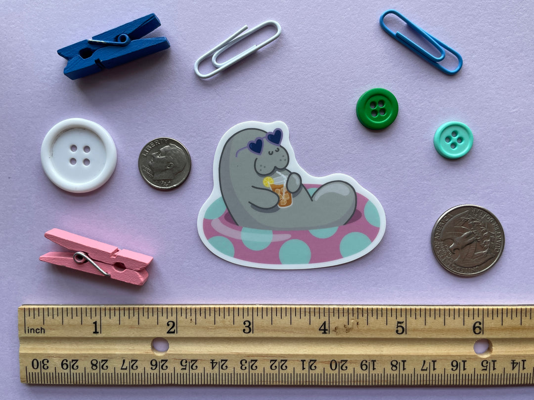 A sticker of a grey cartoon manatee with purple heart-shaped sunglasses holding a glass of iced tea lounging in a pink inner tube with green polka dots is shown on a purple background surrounded by small objects.