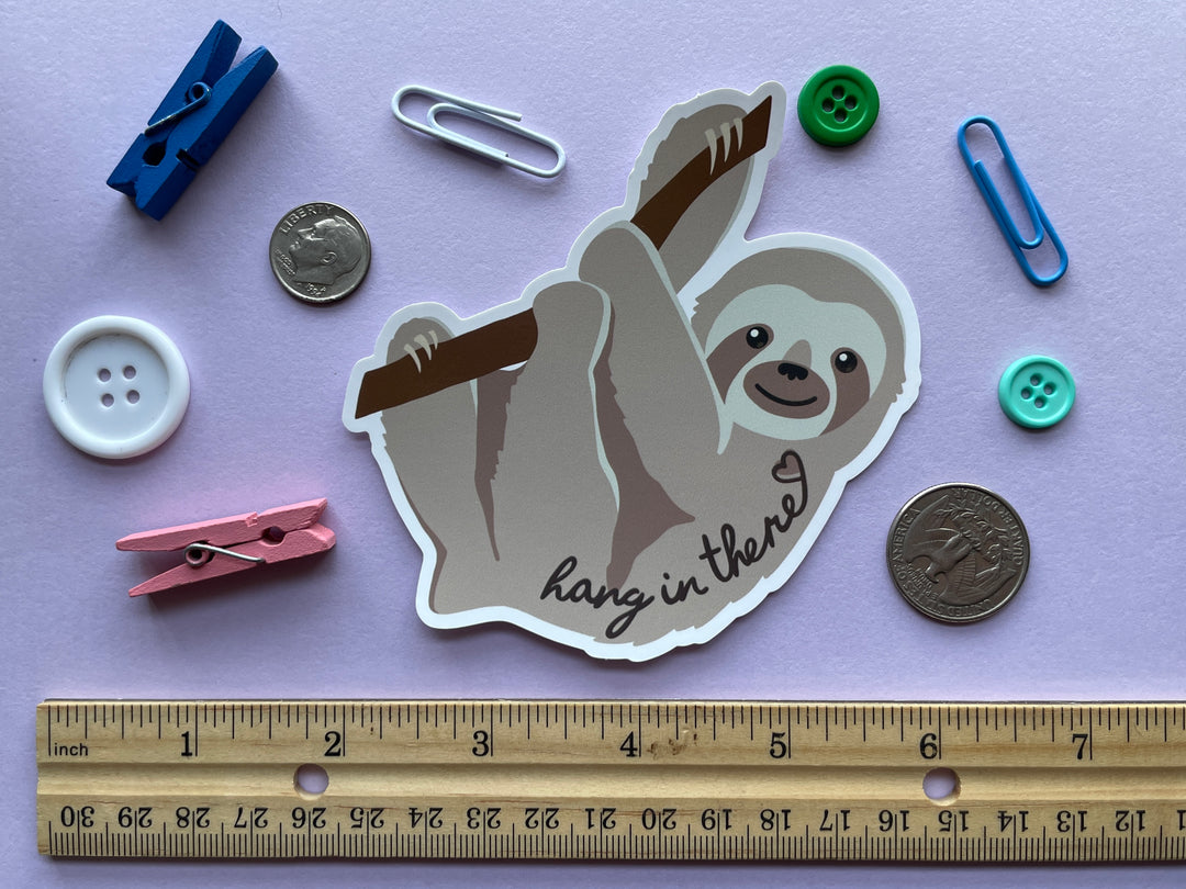 A static cling of a light brown sloth in profile hanging from a brand with the text &quot;hang in there&quot; and a heart over top is shown on a purple background surrounded by small objects.