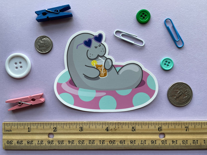 A static cling of a grey cartoon manatee with purple heart-shaped sunglasses holding a glass of iced tea lounging in a pink inner tube with green polka dots is shown on a purple background surrounded by small objects.