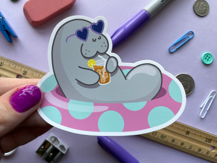 A static cling of a grey cartoon manatee with purple heart-shaped sunglasses holding a glass of iced tea lounging in a pink inner tube with green polka dots is shown held over a purple background covered in objects.