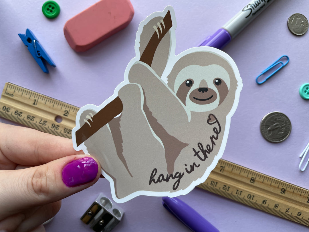 A static cling of a light brown sloth in profile hanging from a brand with the text &quot;hang in there&quot; and a heart over top is held over a purple background covered in objects.