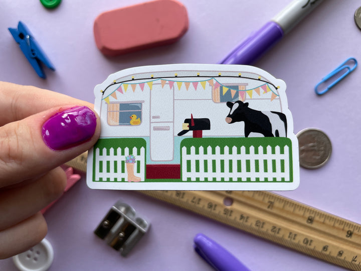 A magnet of the caravan from the TV show Taskmaster with a cow in the front yard and duck in the window is held over a purple background covered in objects.