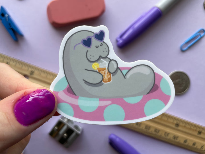 A sticker of a grey cartoon manatee with purple heart-shaped sunglasses holding a glass of iced tea lounging in a pink inner tube with green polka dots is shown held over a purple background covered in objects.