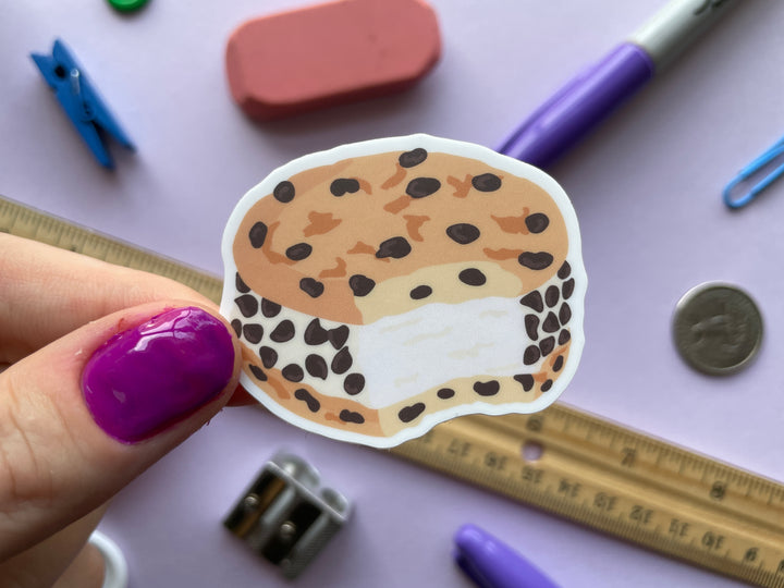 A sticker of a chocolate chip cookie ice cream sandwich is held above a purple background.