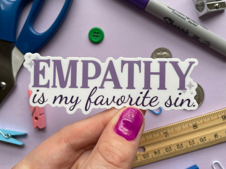 A sticker of purple text that reads &quot;empathy is my favorite sin&quot; is held above a purple background covered in small objects.