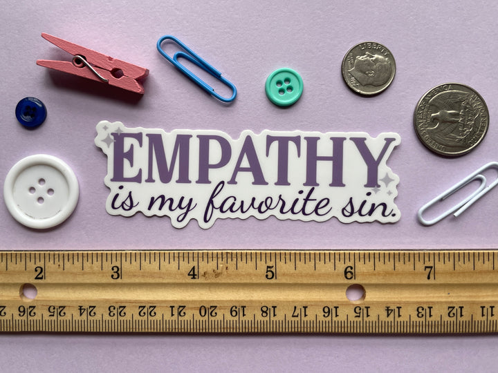 A sticker of purple text that reads &quot;empathy is my favorite sin&quot; is shown on a purple background covered in small objects.