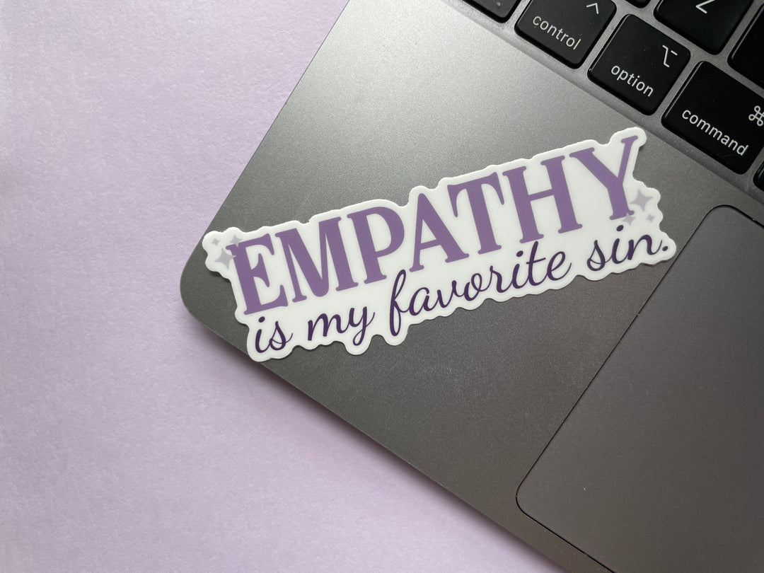 A sticker of purple text that reads &quot;empathy is my favorite sin&quot; is shown on a silver laptop resting on a purple background.
