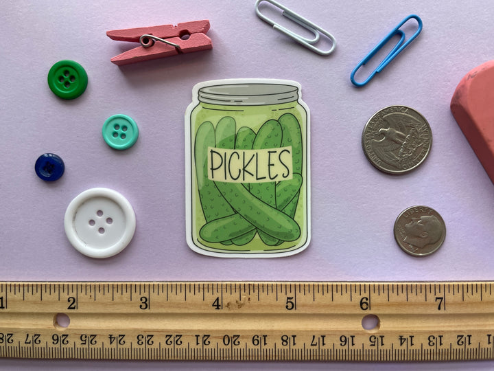 A sticker of a cartoon drawing of a jar of whole dill pickles with a label reading "pickles" is shown on a purple background surrounded by small objects.