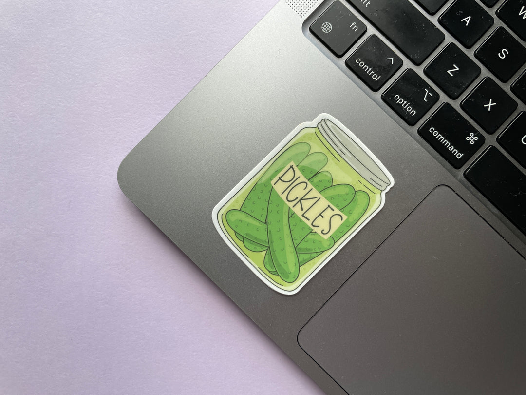 A sticker of a cartoon drawing of a jar of whole dill pickles with a label reading "pickles" is shown on a silver laptop on a purple background.