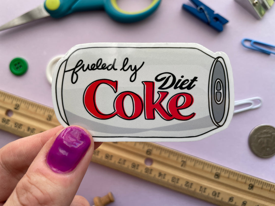 A magnet of a cartoon of a silver can of Diet Coke on its side with the text "fueled by" in the top corner is held over a purple background covered in objects.