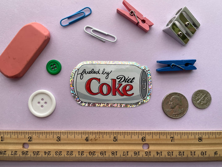A glittery sticker of a cartoon of a silver can of Diet Coke on its side with the text "fueled by" in the top corner is shown on a purple background surrounded by small objects.