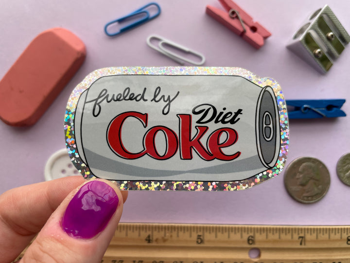A glittery sticker of a cartoon of a silver can of Diet Coke on its side with the text "fueled by" in the top corner is held above a purple background covered in small objects.