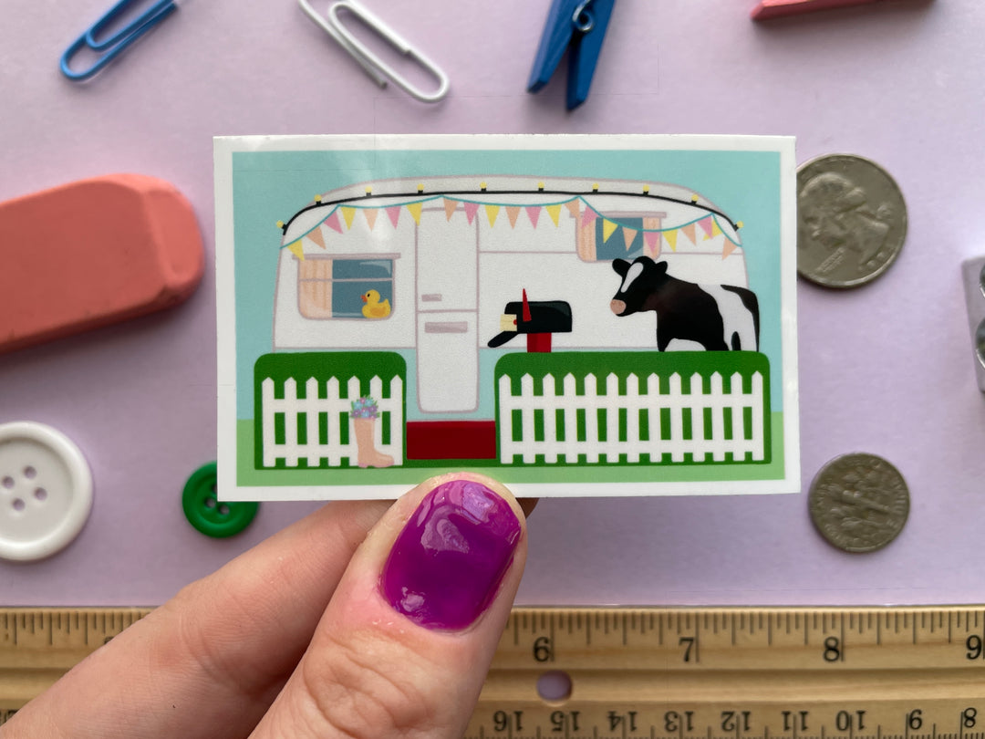 A magnet of the caravan from the TV show Taskmaster with a cow in the front yard and duck in the window is held over a purple background covered in objects.
