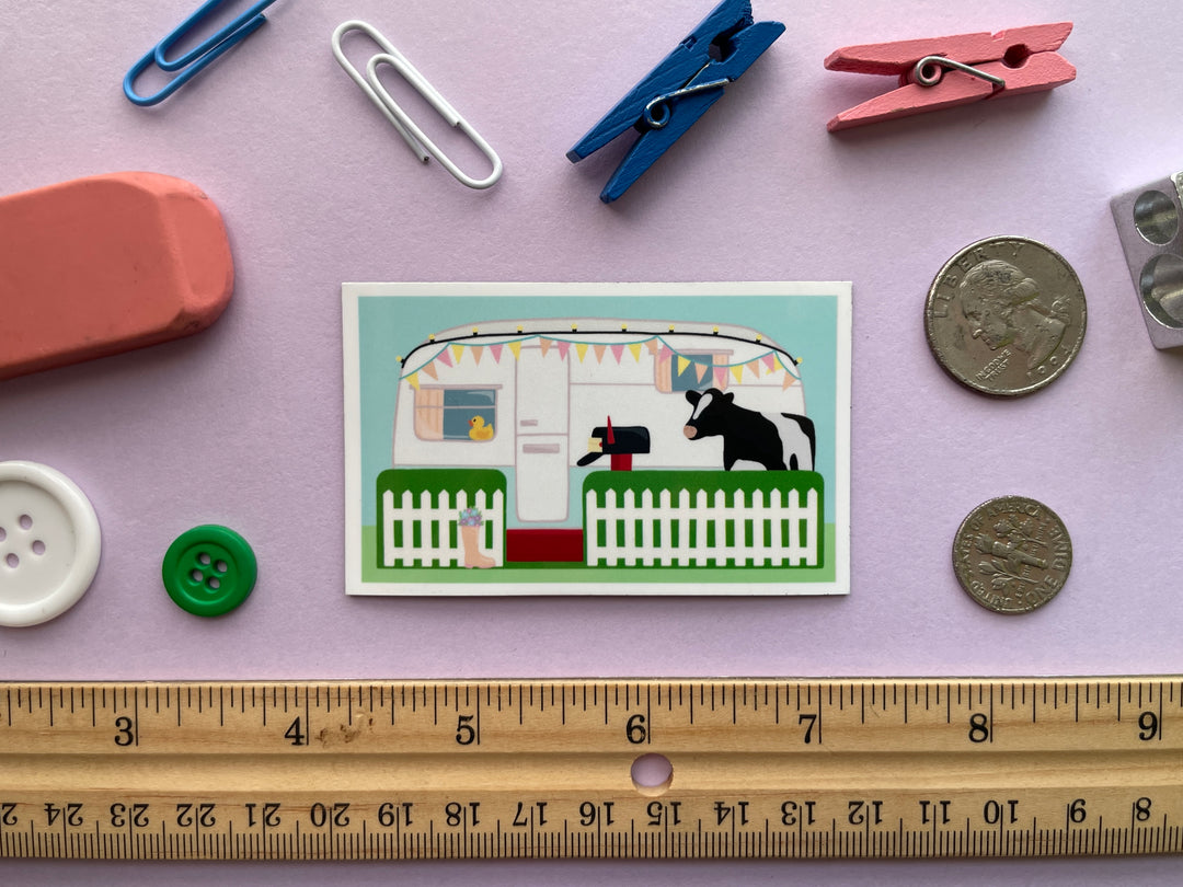 A magnet of the caravan from the TV show Taskmaster with a cow in the front yard and duck in the window is shown on a purple background surrounded by objects.